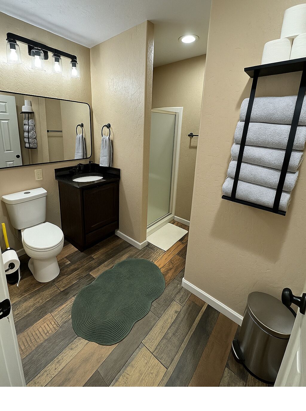 A bathroom with a toilet, sink, mirror, towel rack, shower, and trash can.