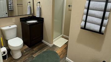 A bathroom with a toilet, sink, mirror, towel rack, shower, and trash can.