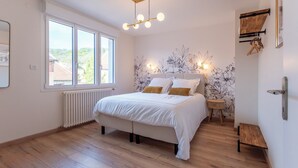 Een slaapkamer met een groot bed, een raam, een radiator, een houten vloer, een wandlamp en een klein houten bankje.