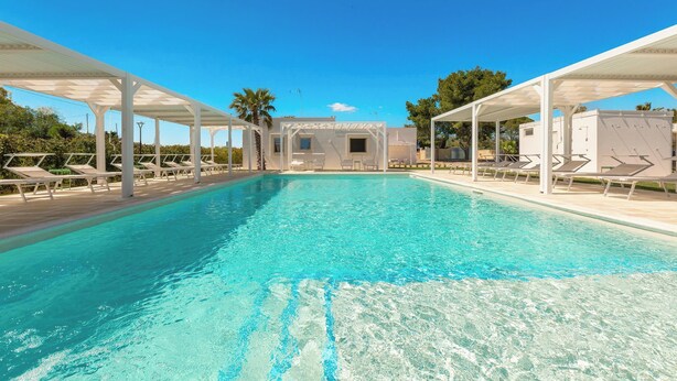 A clear blue pool with white sunshades and lounge chairs, surrounded by a covered area with a view of a building in the background. - B&B Giorò | Camera tripla (Galatone)