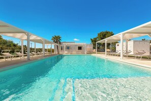 Une piscine d’un bleu éclatant, agrémentée de parasols blancs et de chaises longues, entourée d’une zone abritée d’où l’on peut admirer un bâtiment en arrière-plan.
