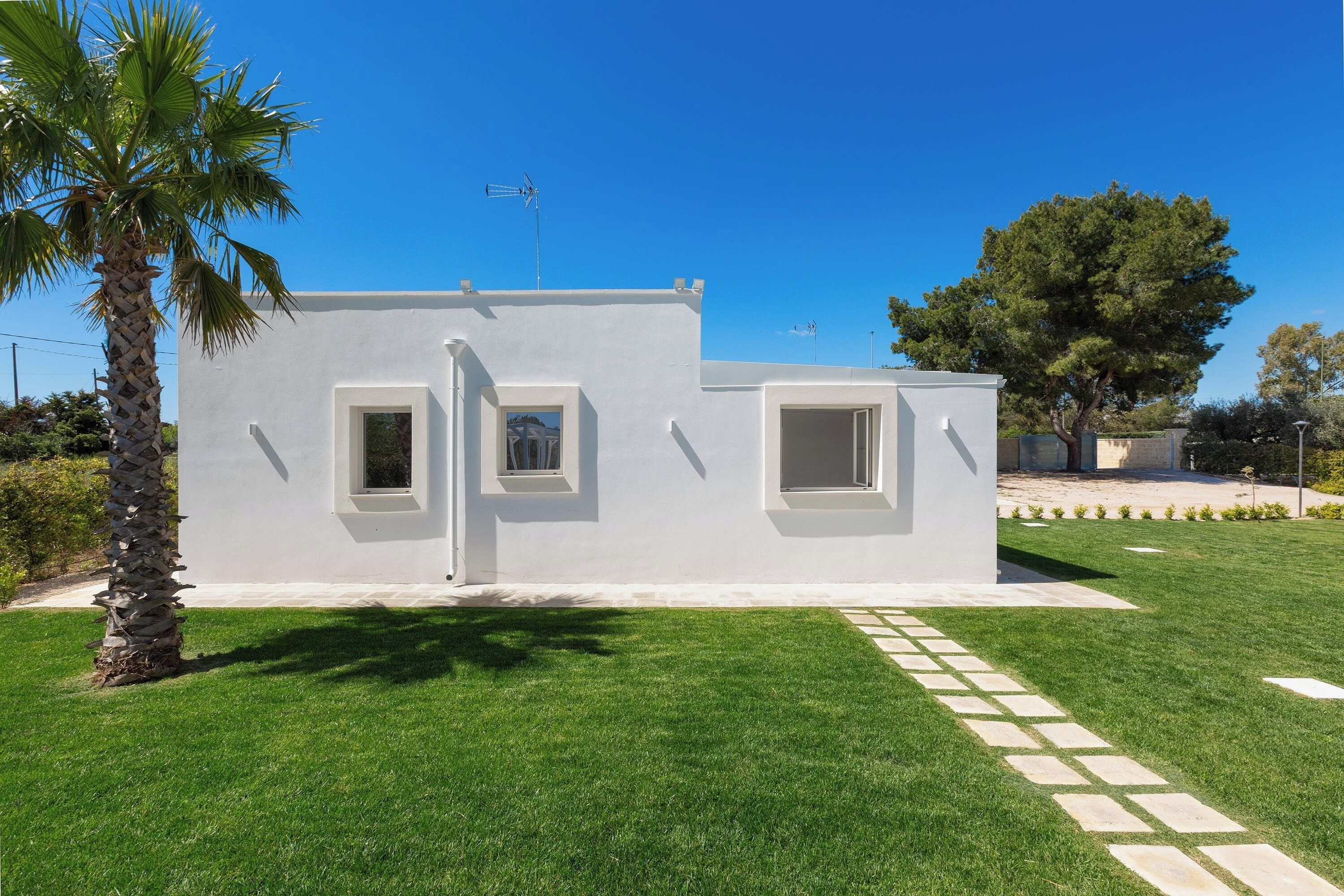 Un edificio moderno blanco con dos ventanas pequeñas y una puerta, rodeado de un césped cuidado y una palmera.