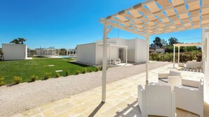 Un área de estar techada al aire libre con mobiliario de mimbre blanco, vista despejada a una piscina y un césped bien cuidado.