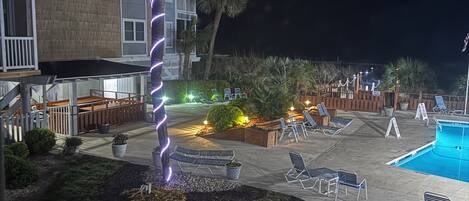 A nighttime view of a hotel pool area with lounge chairs, a palm tree, and a building with balconies.