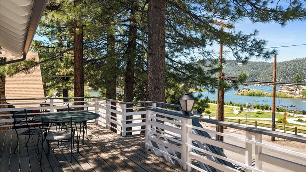A wooden deck with a table and chairs, overlooking a lake with a white fence and trees.