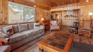 A cozy cabin interior with a wooden ceiling, a sofa with a 'HOME' pillow, a coffee table, and a rocking chair.