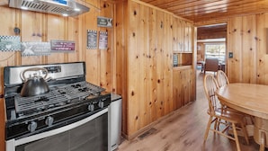 A wooden-paneled kitchen with a gas stove, oven, and a kettle. There are signs on the wall and a dining table with chairs.