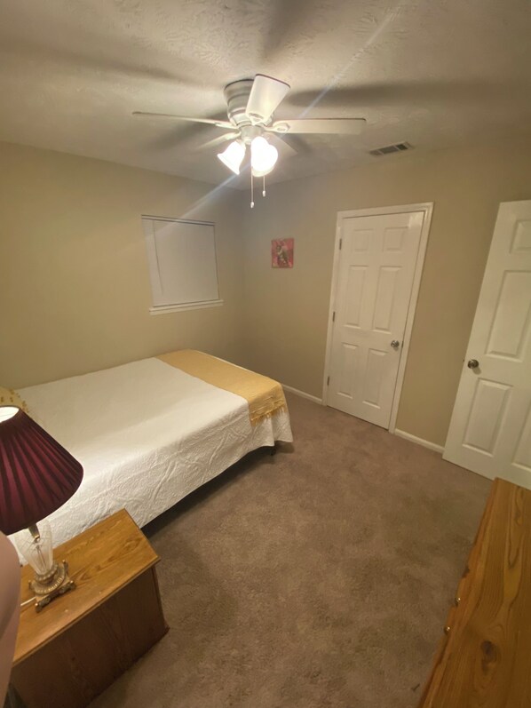 Une chambre à coucher avec un lit, un ventilateur de plafond, une porte et une petite commode en bois.
