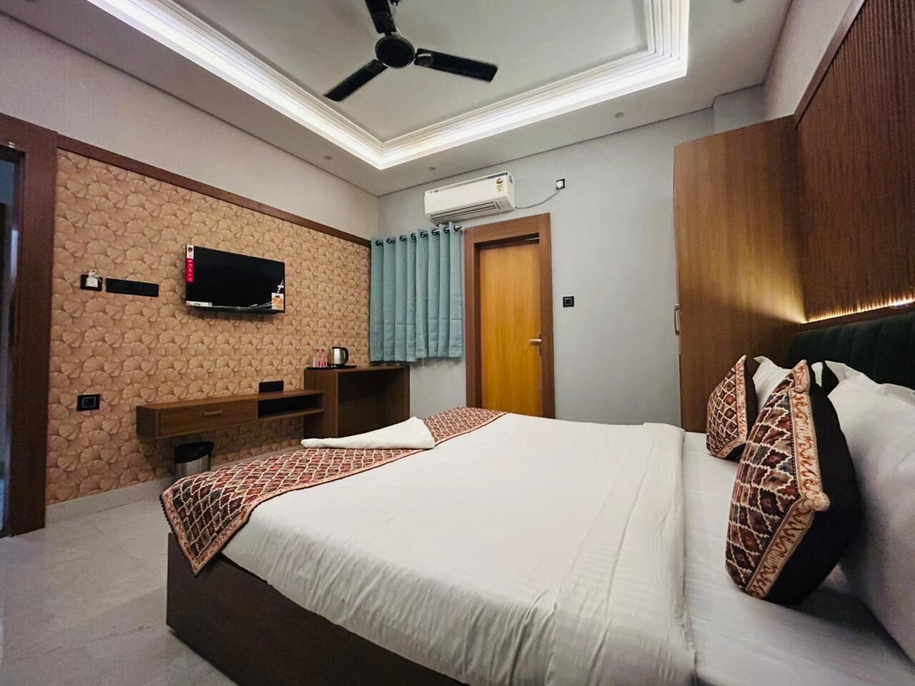 A hotel room with a bed, a ceiling fan, a TV mounted on the wall, and a wooden wardrobe.