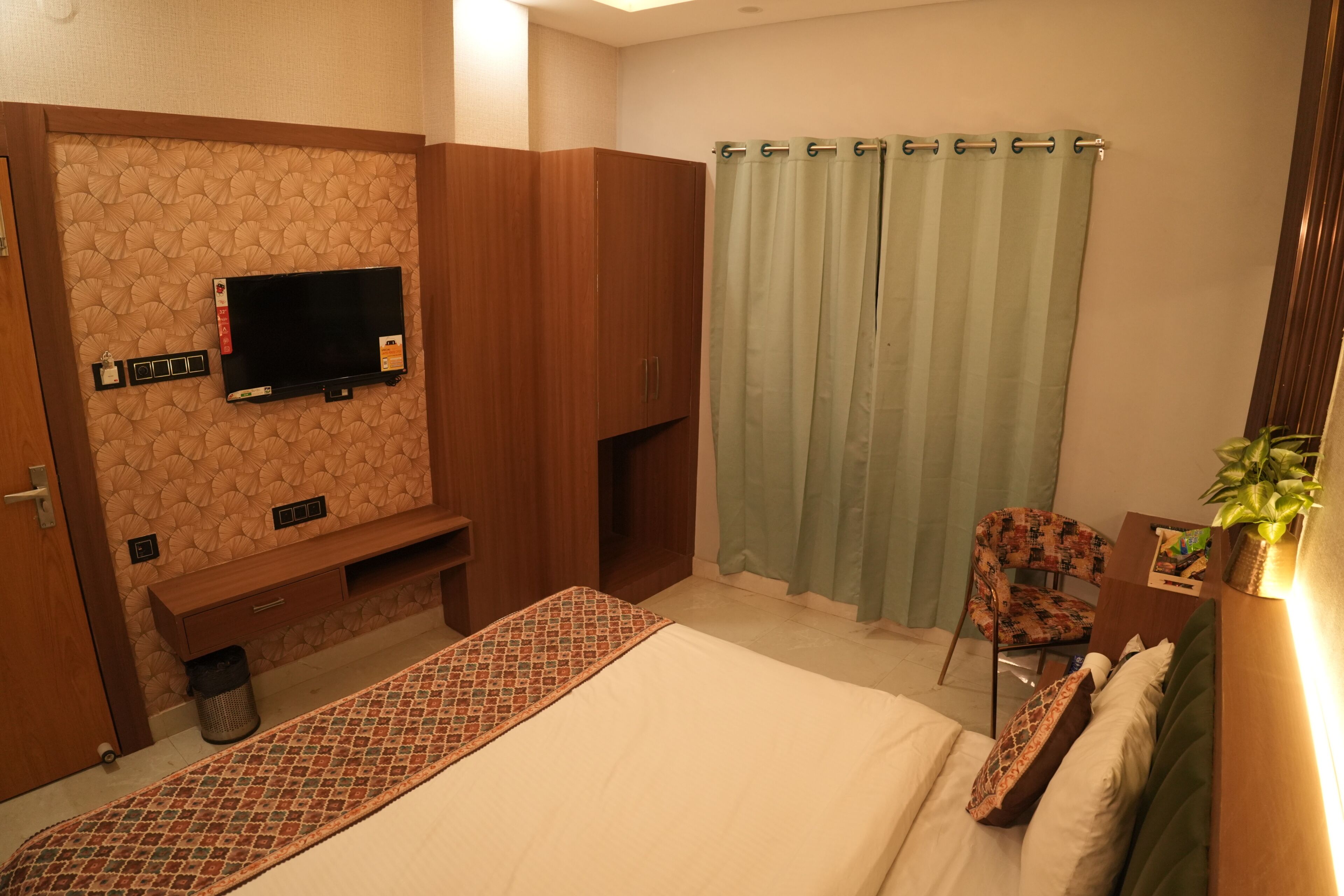 A hotel room with a bed, a TV mounted on the wall, a small seating area, and a curtain.