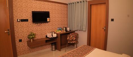 A hotel room with a bed, a TV, a desk with a chair, a plant, and an air conditioning unit.