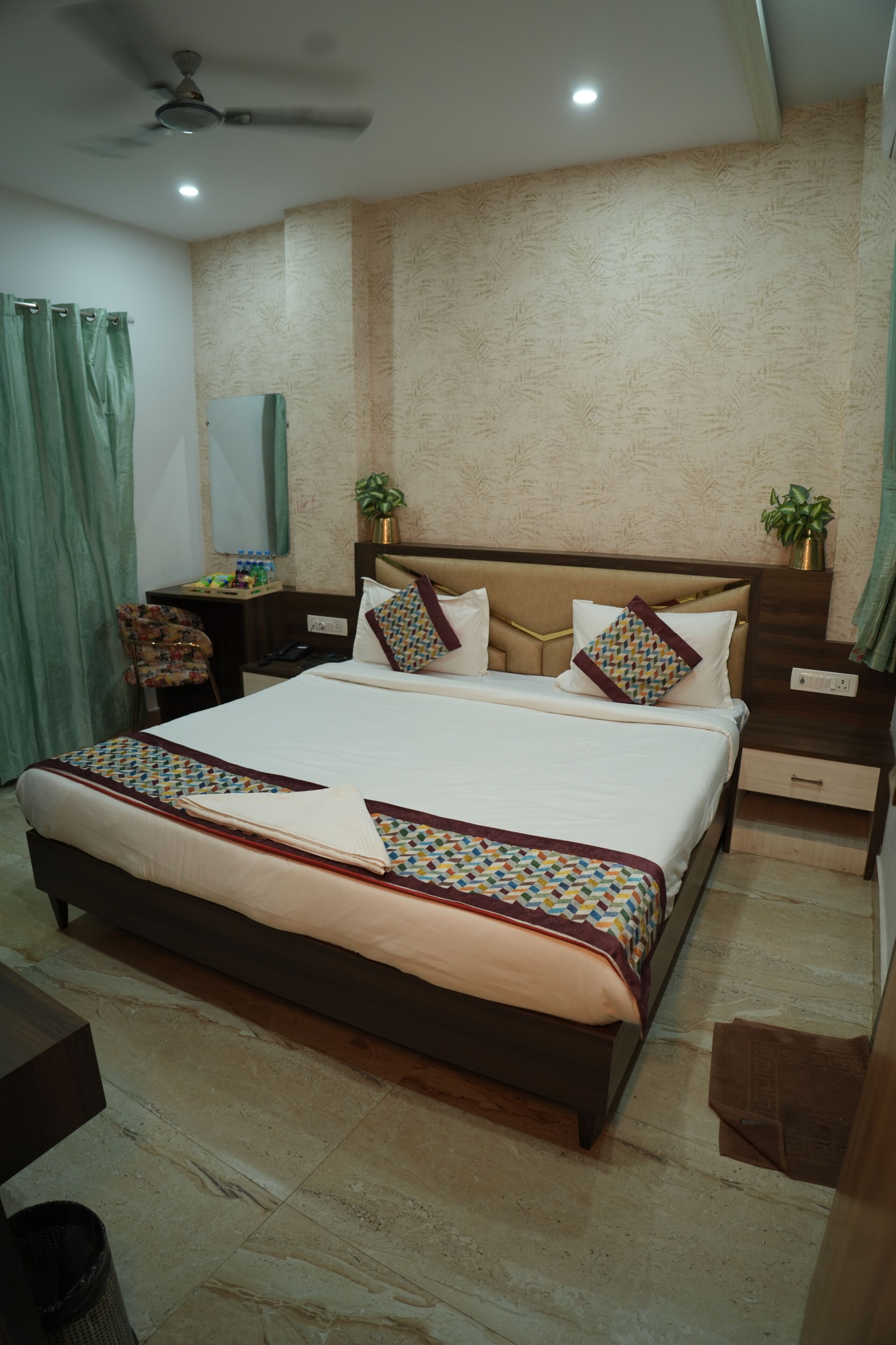 A hotel room with a large bed, a desk, a chair, a nightstand, a ceiling fan, and a wall-mounted mirror.