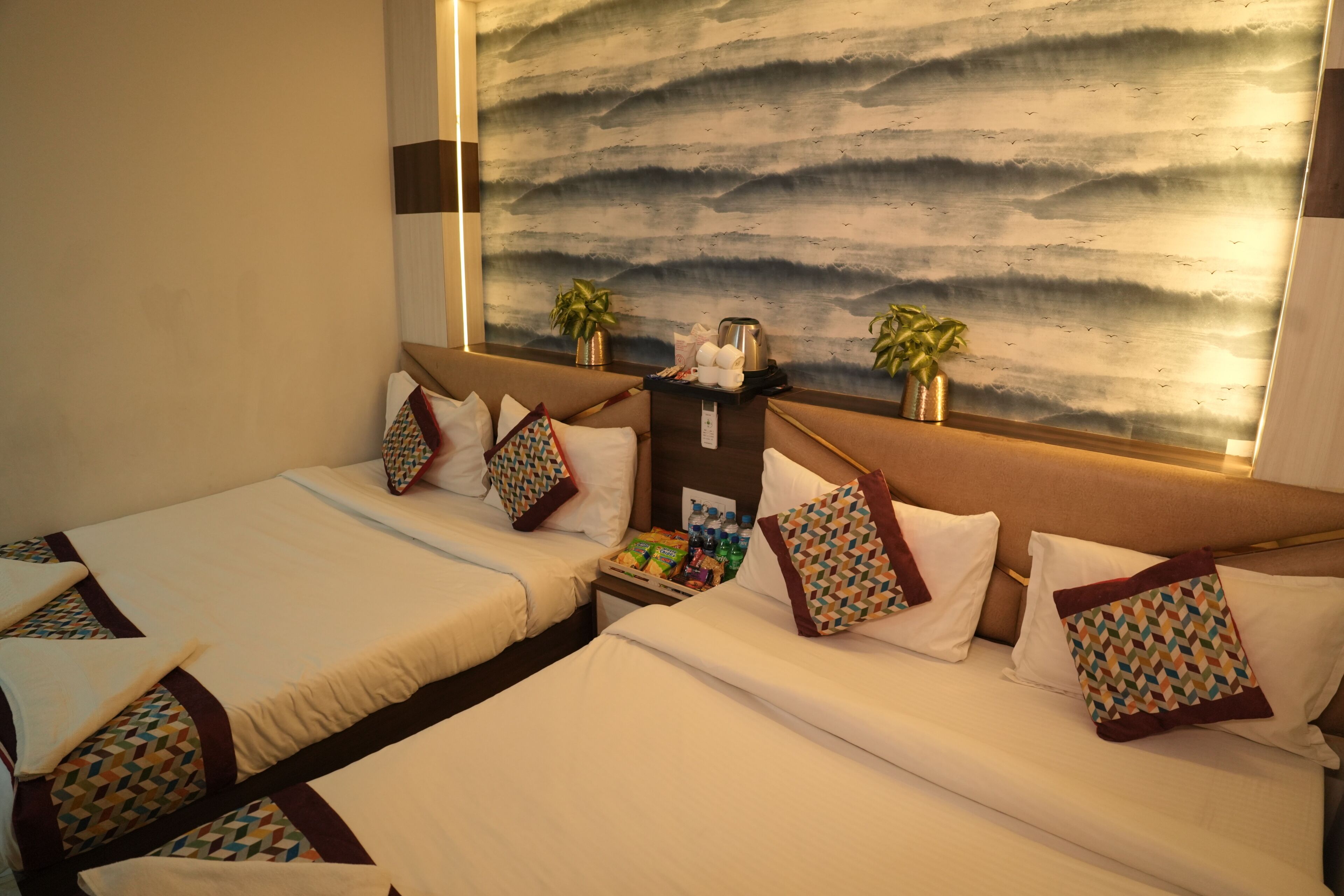 A hotel room with two beds, a large abstract wall mural, two potted plants, and a small table with a kettle and snacks.
