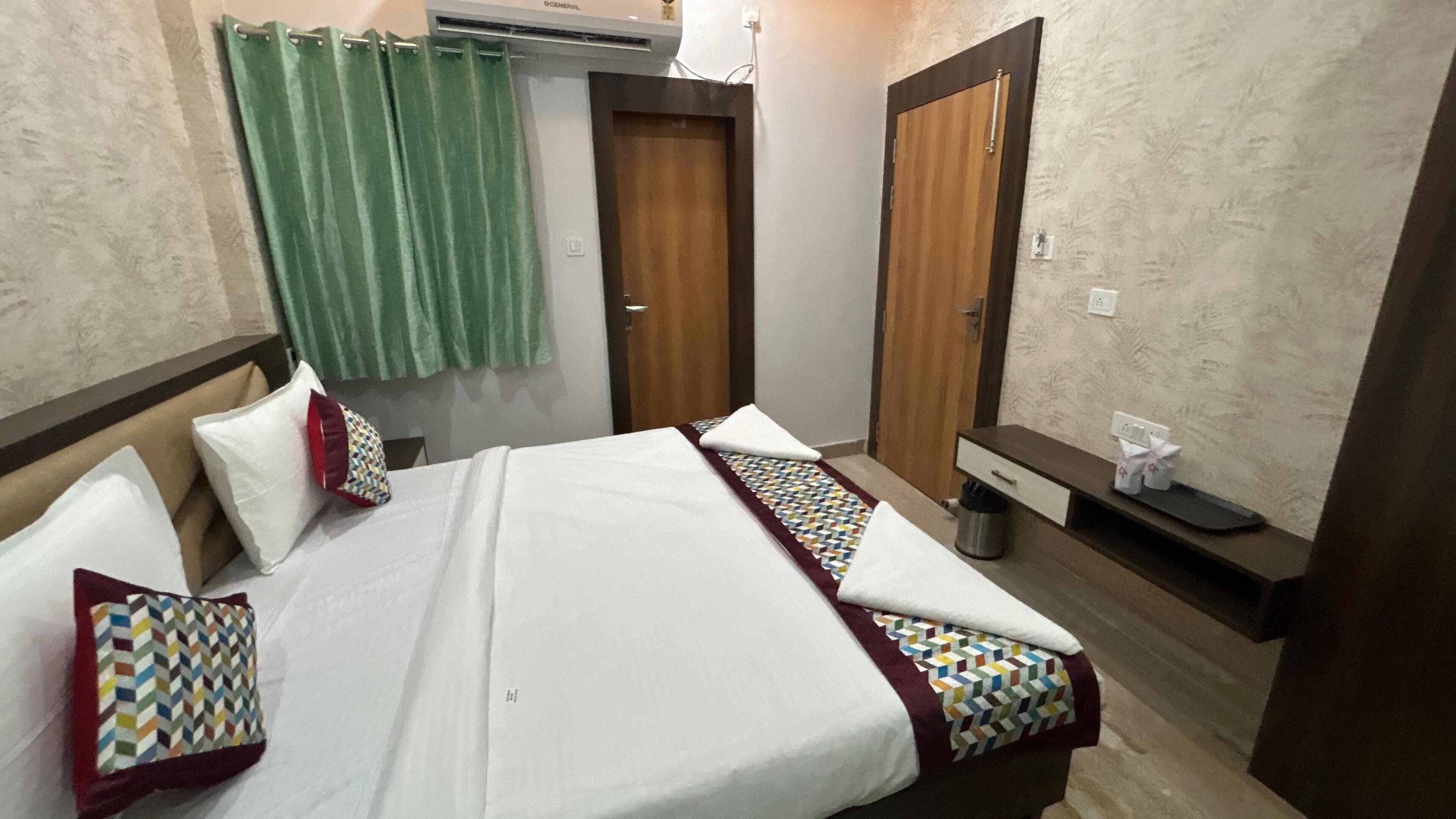 A hotel room with a bed, a small bedside table, a mirror, a green curtain, and a wooden door.