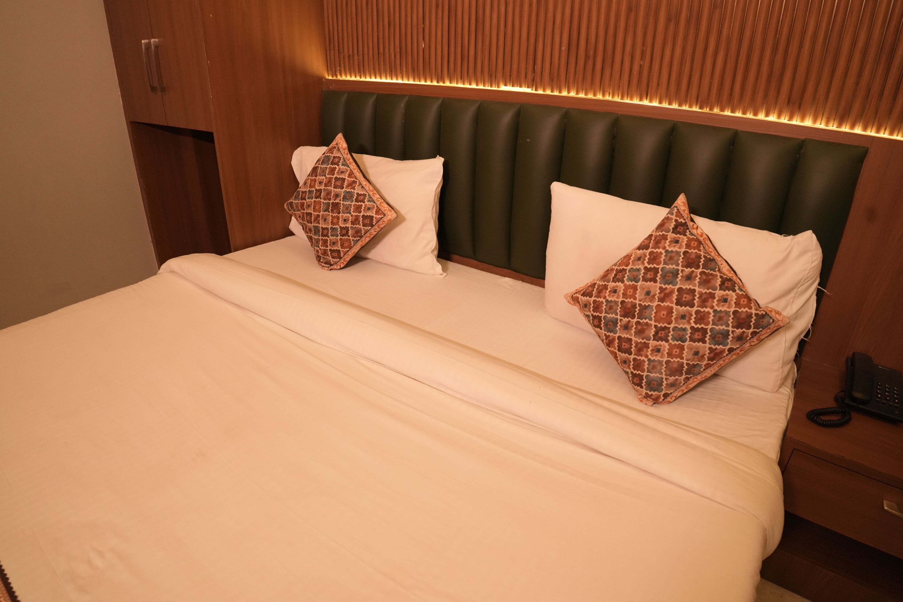 A hotel room with a bed, two patterned pillows, a wooden headboard, and a bedside telephone.