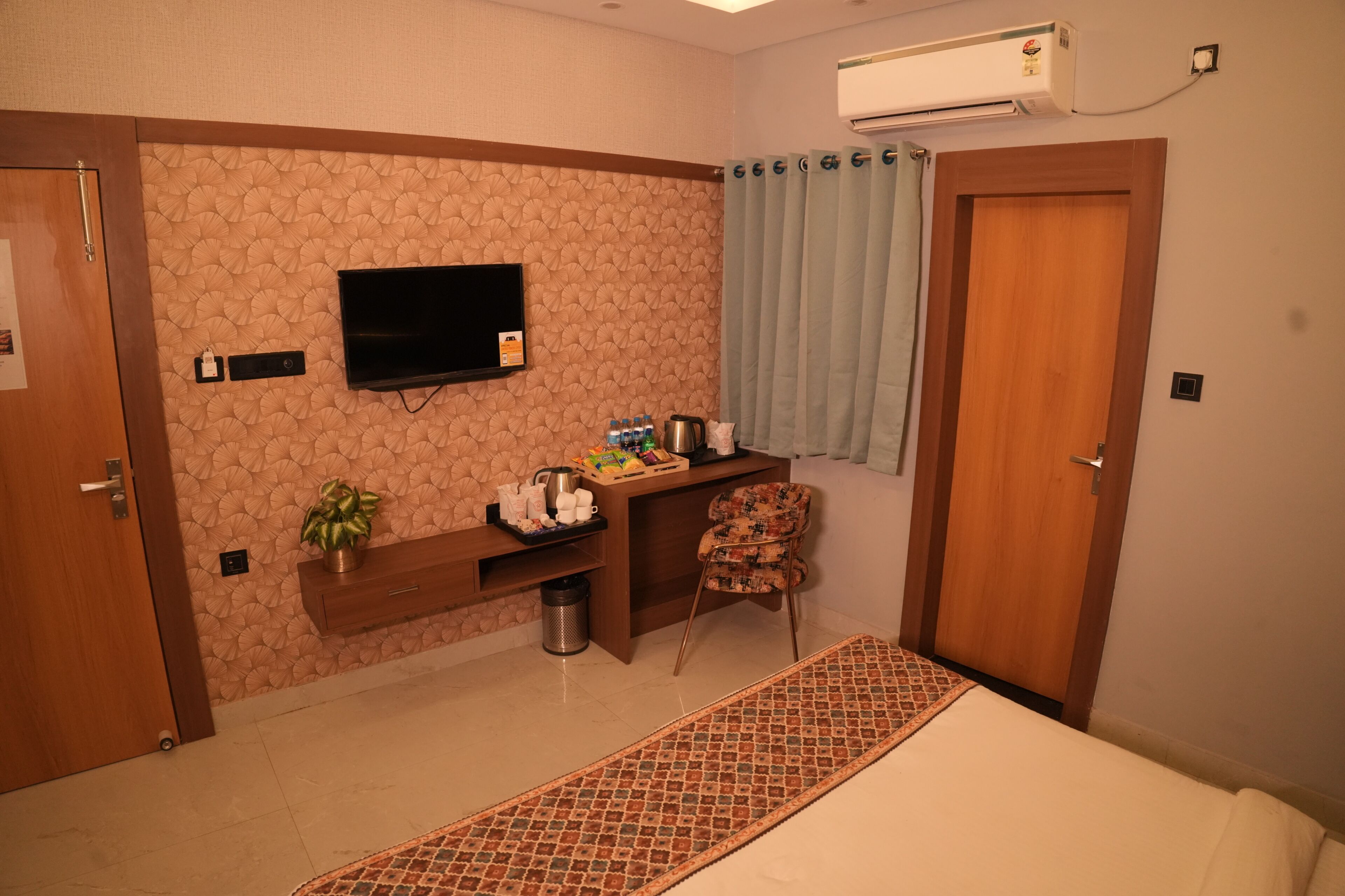 Habitación de hotel con una cama, televisión montada en la pared, escritorio con silla, planta y ventana con cortina.