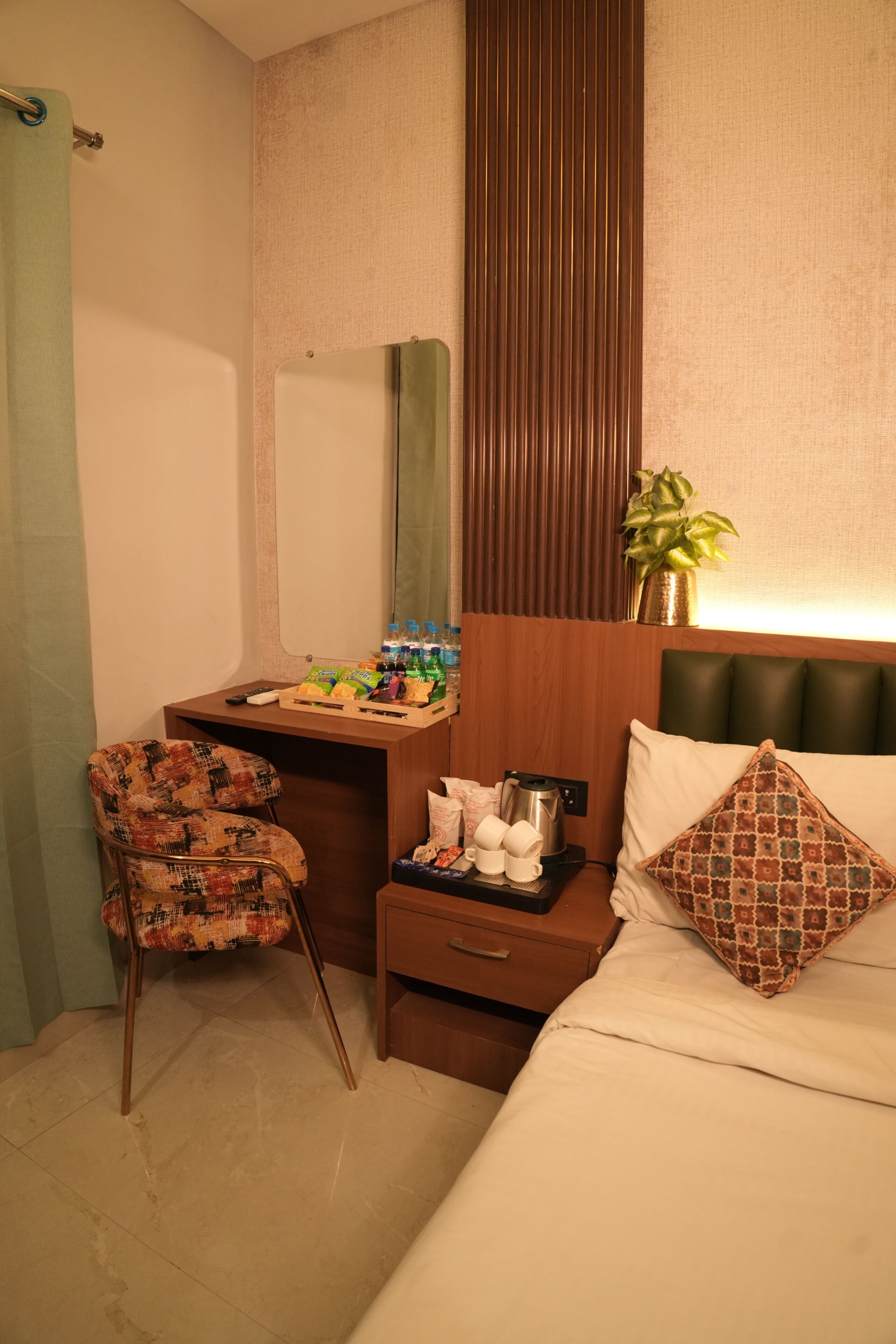 A hotel room with a bed, a desk with a chair, a mirror, a plant, and a tray with snacks.