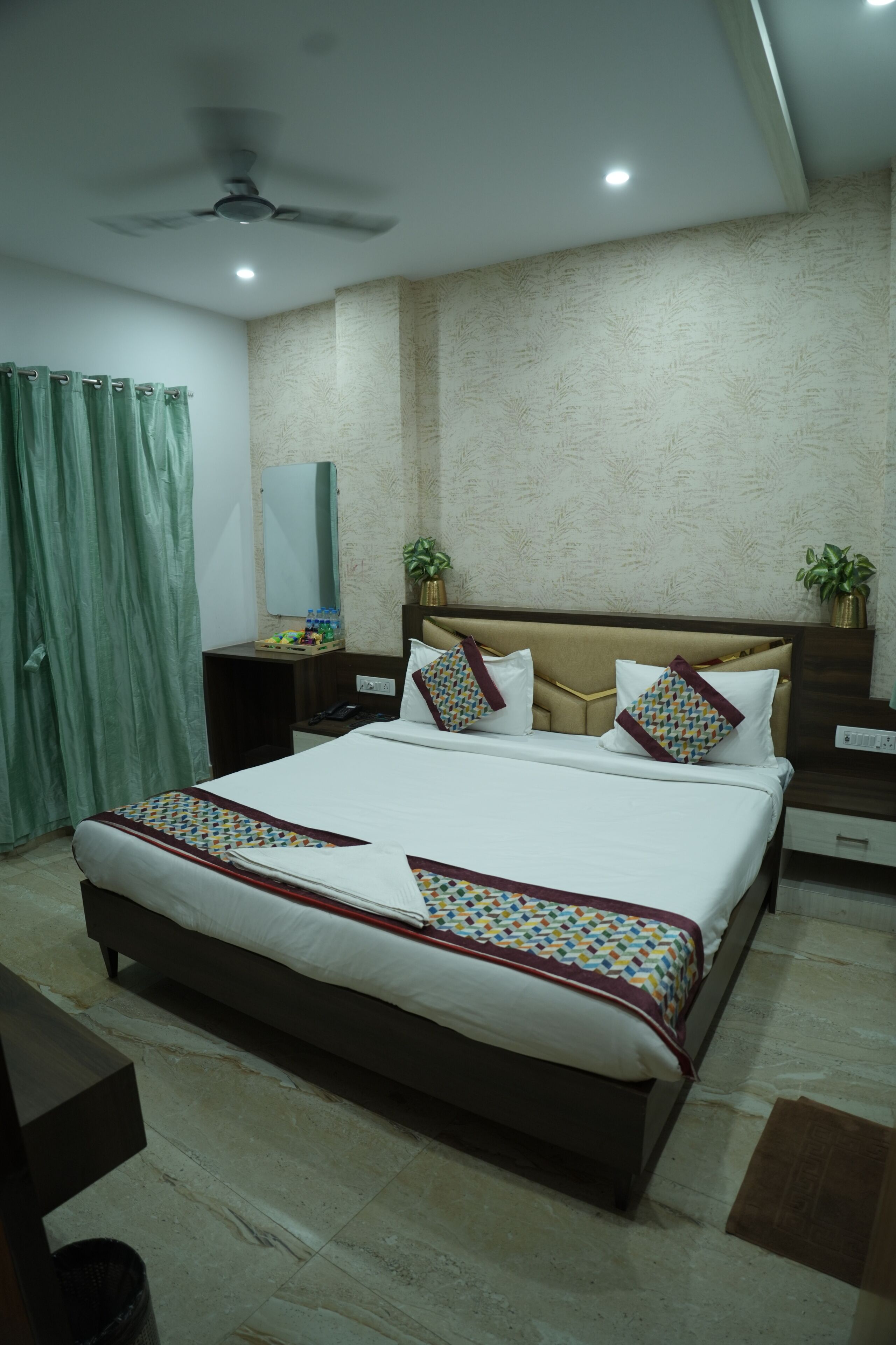 Quarto de hotel com uma cama, um ventilador de teto, uma mesa de cabeceira, um espelho e uma cortina verde.