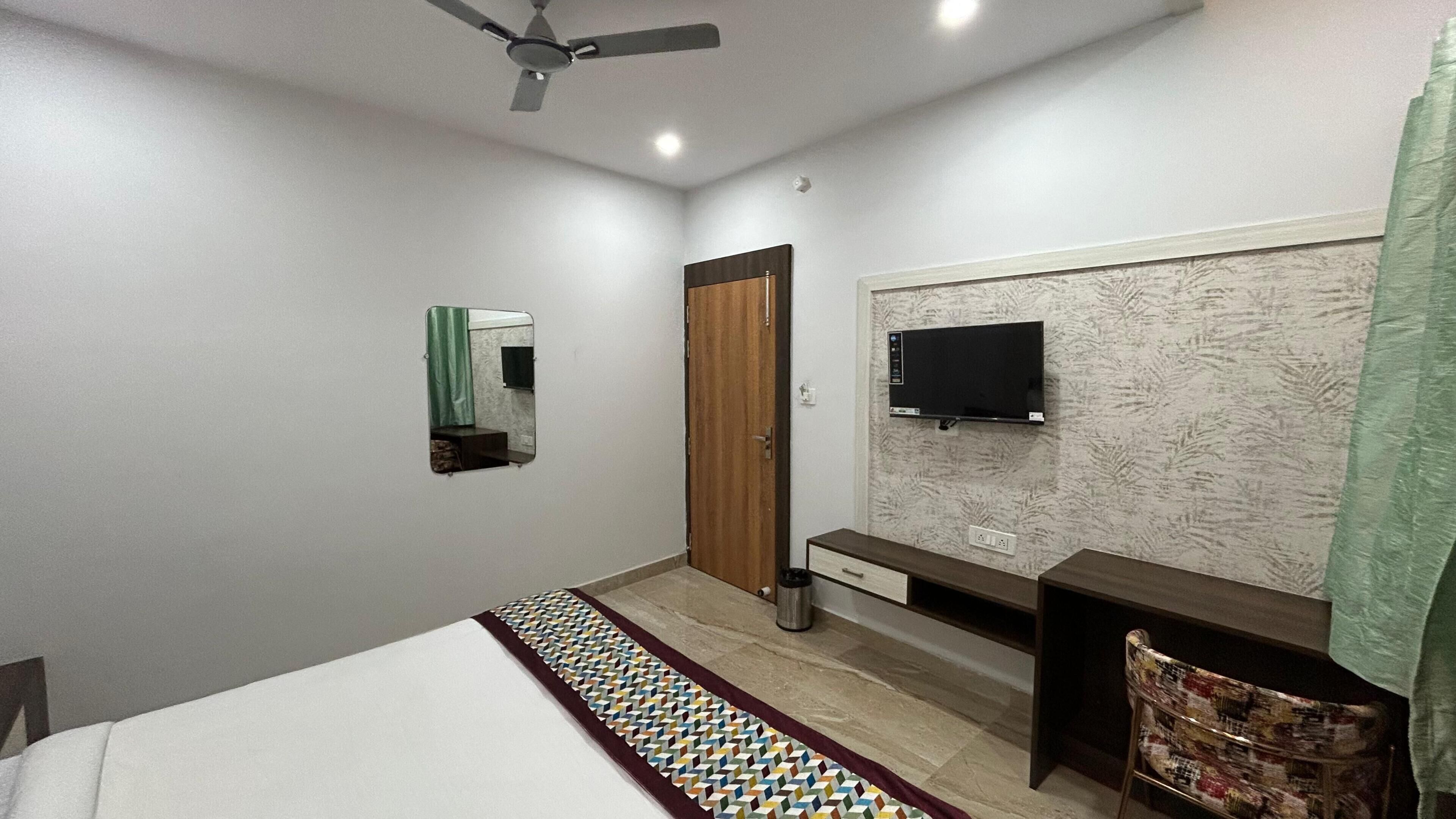 Une chambre à coucher comprenant un lit, un bureau, une chaise, un téléviseur, un ventilateur de plafond et une porte.