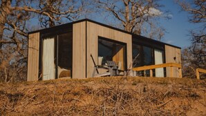 A modern, wooden cabin with large windows, situated on a grassy hill with trees in the background. - Wilding Hotels Scenic Suite (West Tawakoni)