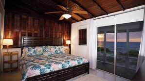 A bedroom with a bed, wooden headboard, ceiling fan, and a view of the ocean through a sliding glass door. - Casa Solemare Oceanfront Cottage (Stake Bay)