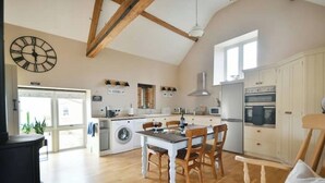 A spacious kitchen with wooden beams, a dining table with chairs, a clock, and a washing machine. - The Stone Barn - Luxury Barn in Rural Wiltshire (Studley)
