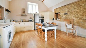 A kitchen with wooden flooring, a dining table with chairs, a washing machine, and a stone wall. - The Stone Barn - Luxury Barn in Rural Wiltshire (Studley)