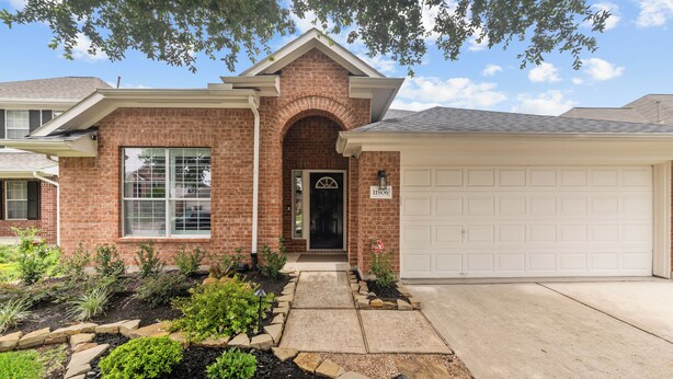 A two-story brick house with a garage, a front door with a decorative arch, and a well-maintained garden with various plants and a pathway leading to the door. - Perfect cottage - Refined Comfort Meets Pet-friendly Backyard Serenity (Tomball)