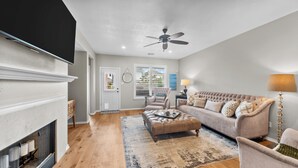 A living room with a fireplace, a sofa, an ottoman, a ceiling fan, and a view of the outside through a window. - Perfect cottage - Refined Comfort Meets Pet-friendly Backyard Serenity (Tomball)