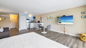 Ein Hotelzimmer mit einem Bett, einem Esstisch mit Stühlen, einer Küchenzeile und einem Fernseher, der eine Strandansicht zeigt.