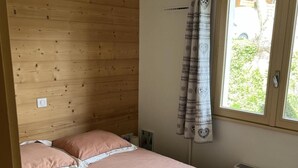 Makuuhuoneessa on puinen sängynpääty, yhden hengen sänky vaaleanpunaisilla vuodevaatteilla, kuvioiduilla verhoilla varustettu ikkuna ja ilmastointilaite.