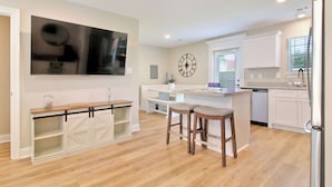 A modern kitchen with a large TV, wooden bar stools, and a clock on the wall. - Steps from the Sand - New 3BR Beach Cottage Near Keesler AFB & Gulf Fun (Biloxi)