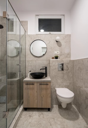 A modern bathroom with a glass-enclosed shower, a wooden vanity with a black sink, a round mirror, and a toilet.