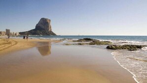 Une plage avec une formation rocheuse imposante en arrière-plan, un rivage sablonneux et quelques personnes qui se promènent.