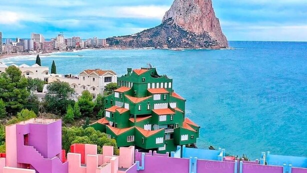 Un paysage urbain côtier avec un imposant bâtiment vert doté de nombreux balcons, une colline rocheuse en arrière-plan et une mer bleue et limpide.