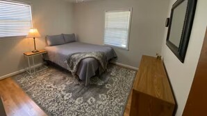 A bedroom with a bed, two windows with blinds, a ceiling fan, a bedside table with a lamp, and a wooden nightstand.