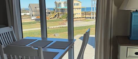 Uma sala de jantar com mesa e cadeiras, uma luminária e vista para a praia e casas através de grandes janelas.