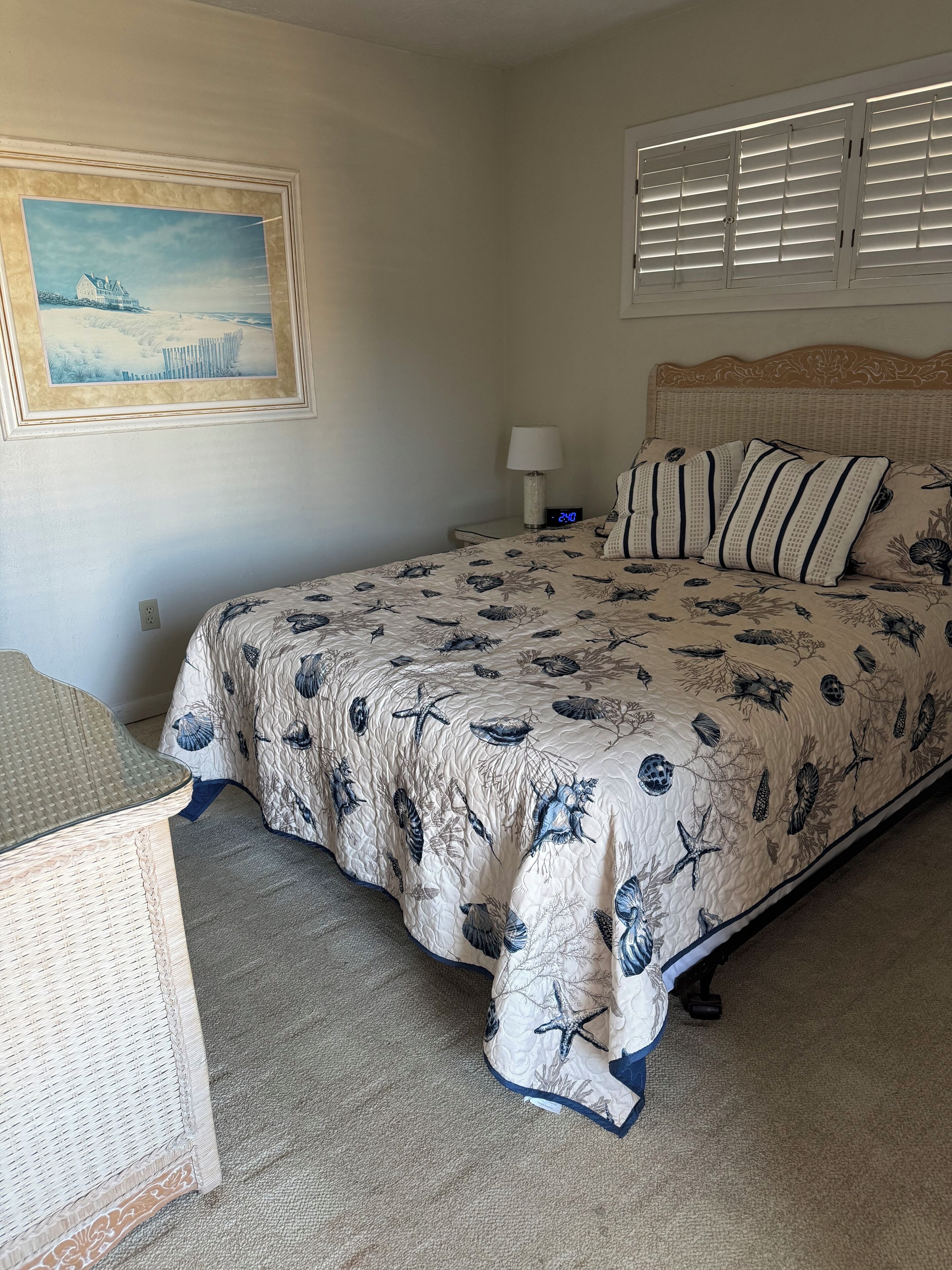 A bedroom with a bed, a nightstand, a lamp, a digital clock, a framed picture on the wall, and a window with blinds.