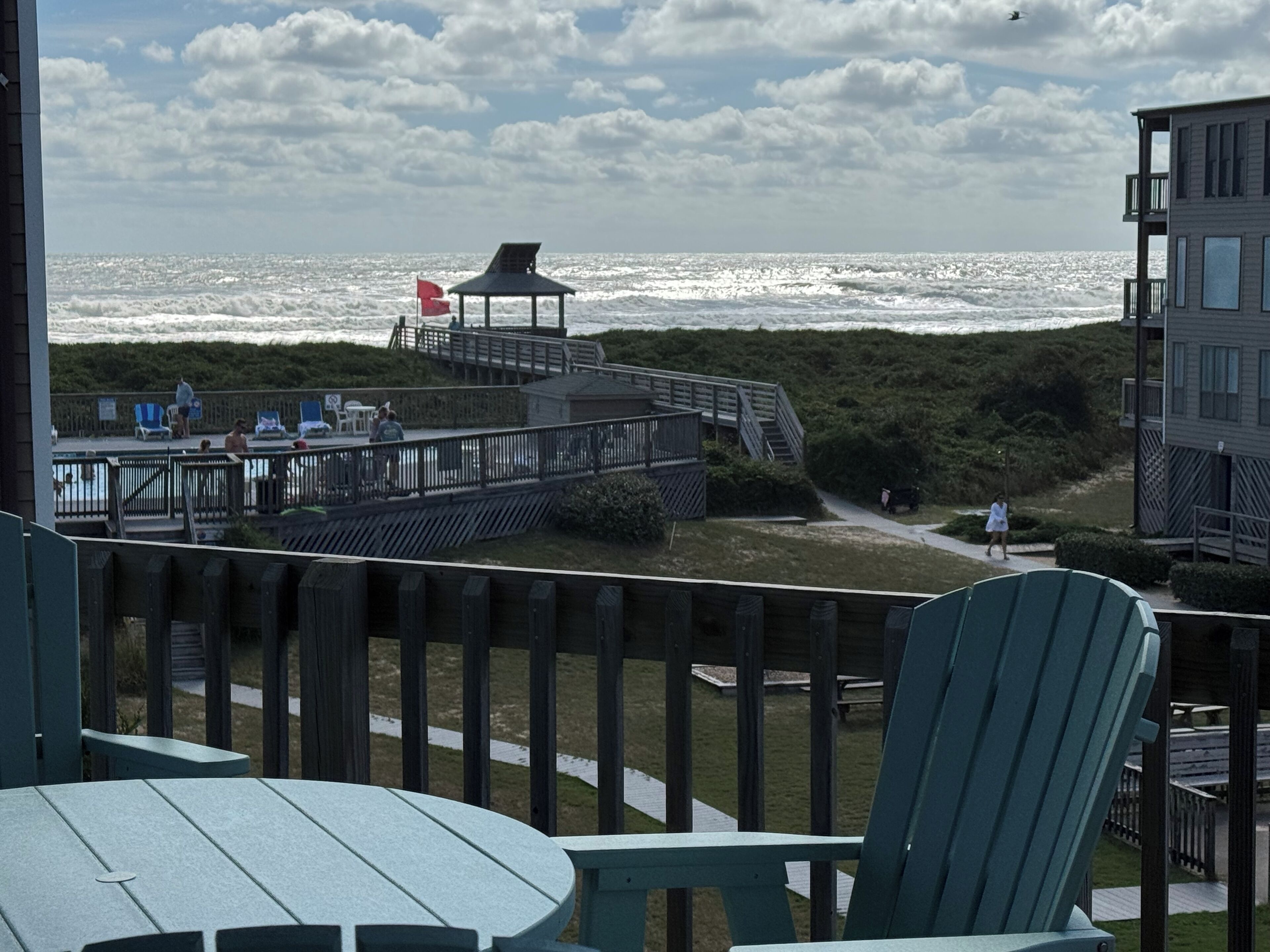A beach view from a balcony with a wooden deck, a gazebo, and a pool area with lounge chairs.