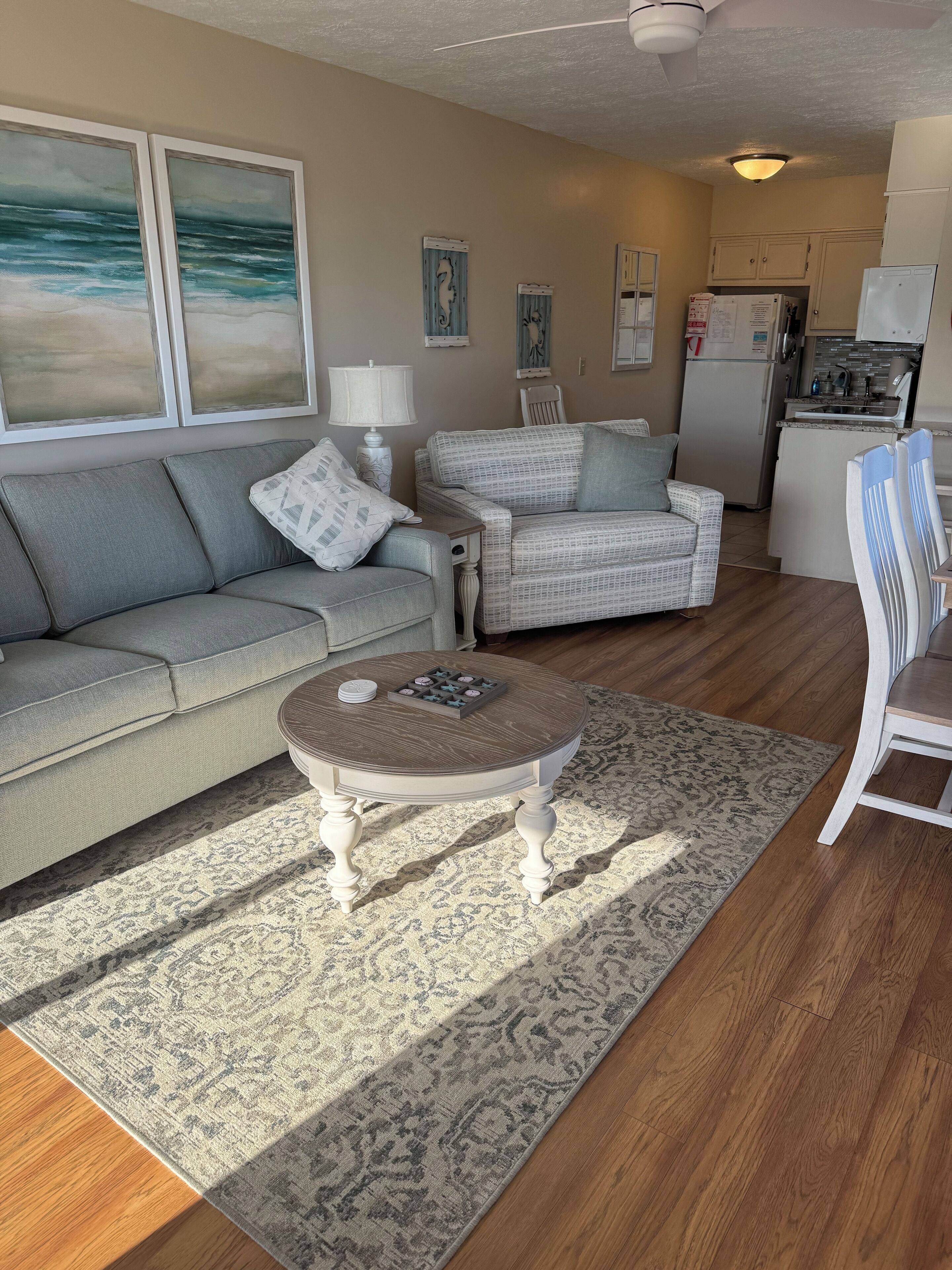 Un salon comprenant un canapé, deux fauteuils, une table basse et un tapis. Il y a également un coin cuisine avec un réfrigérateur et des placards.
