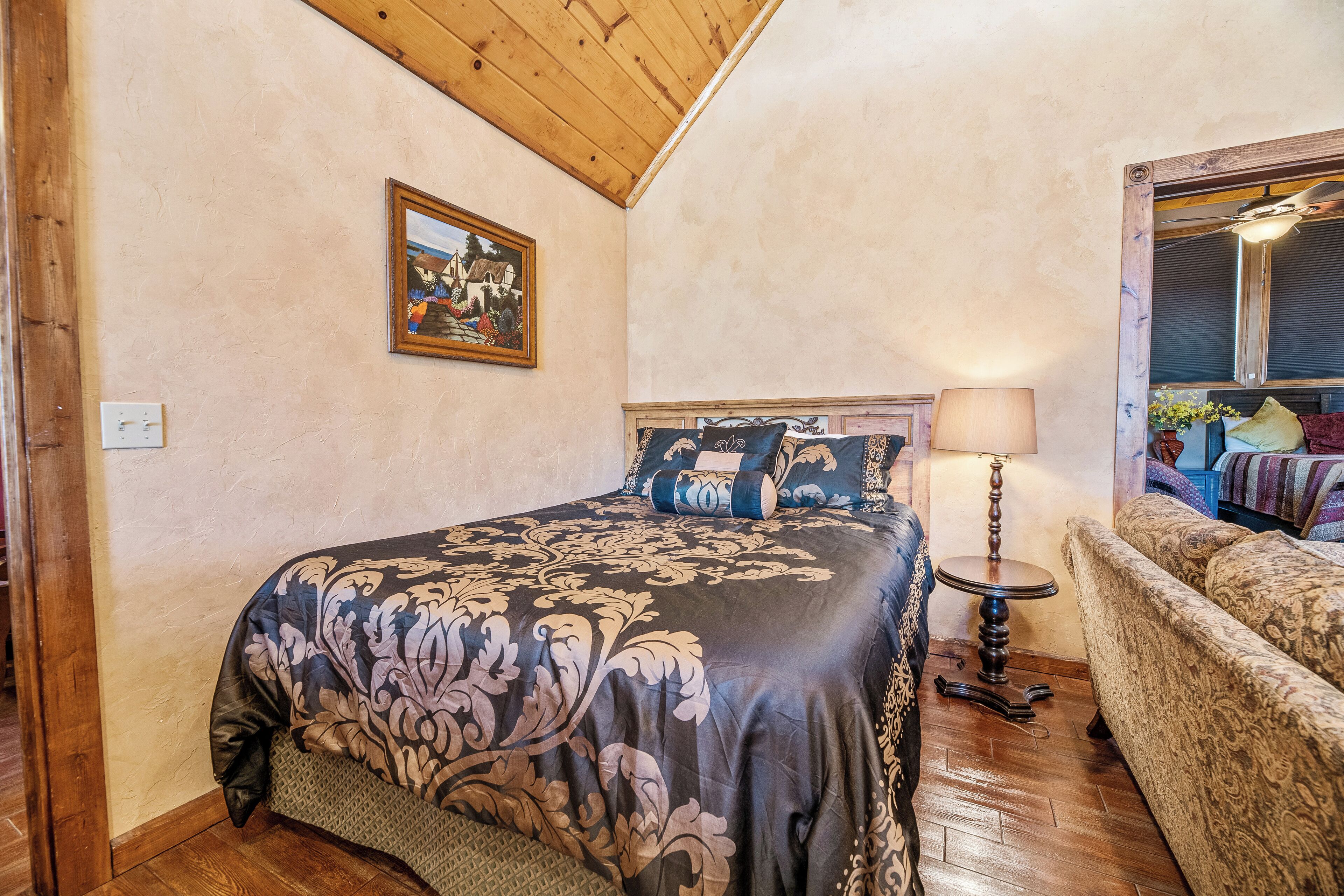 A bedroom with a large bed, a wooden ceiling, a framed picture on the wall, a bedside table with a lamp, and a sofa with patterned cushions.