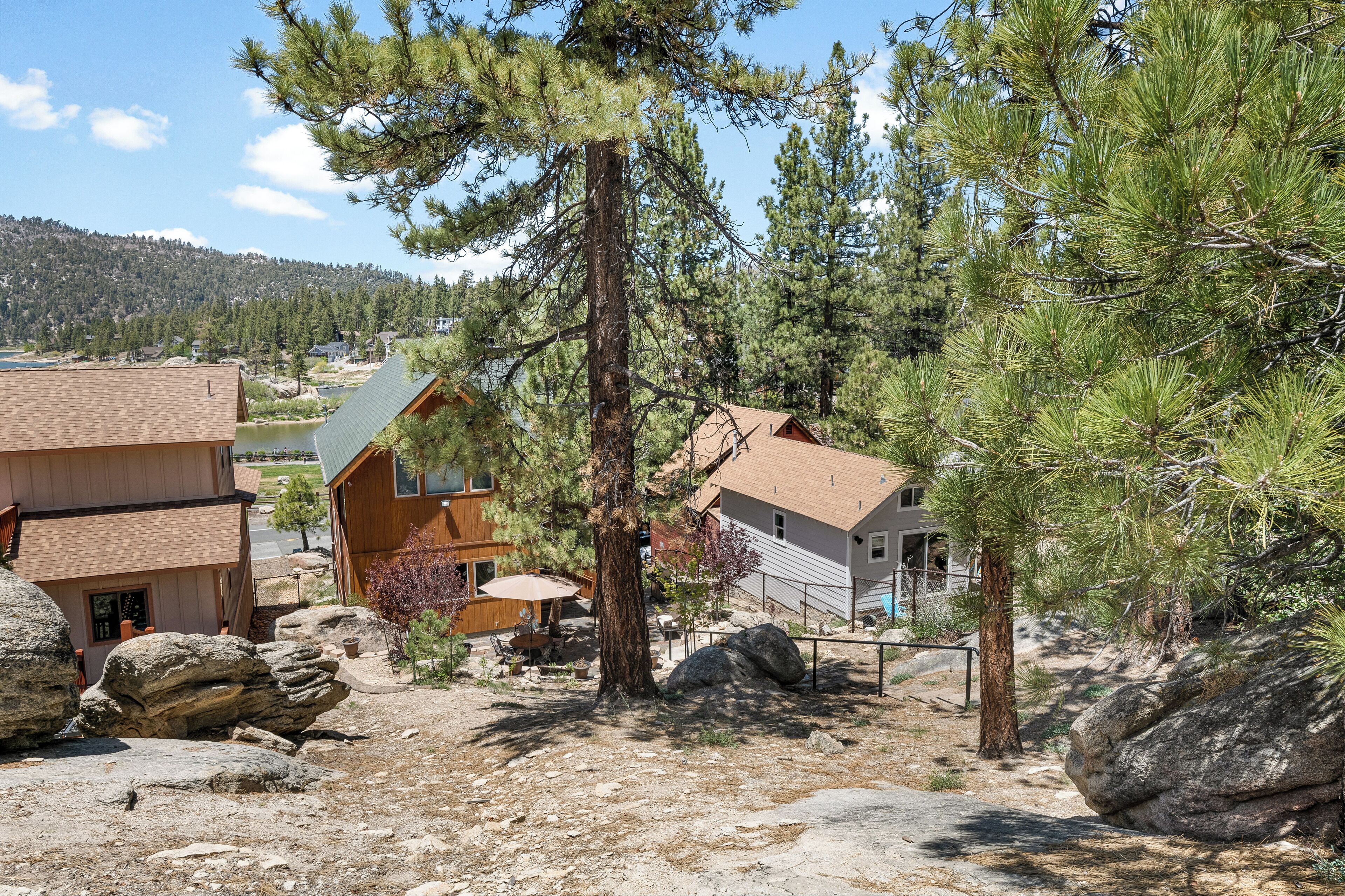 A cabin with a wooden exterior, surrounded by large rocks and pine trees, with a view of a lake and mountains in the background.