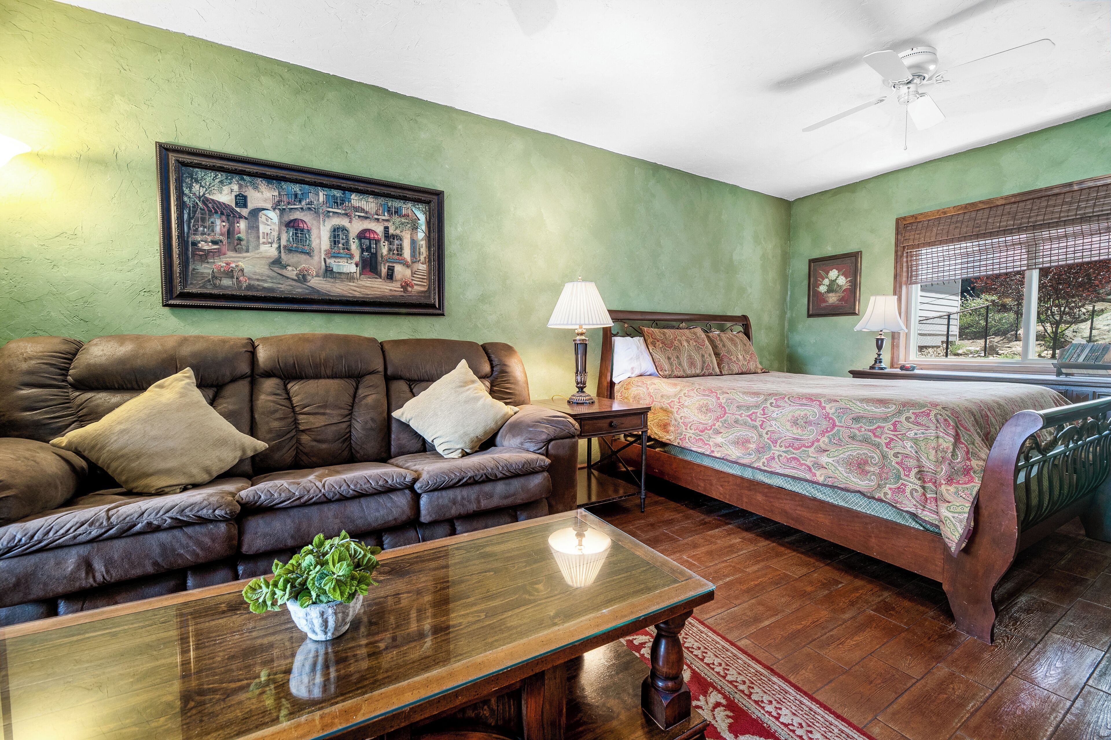 Una stanza con un divano in pelle marrone, un tavolo di legno con una pianta, un letto con copripiumino fantasia, un comodino con una lampada e un quadro appeso al muro.