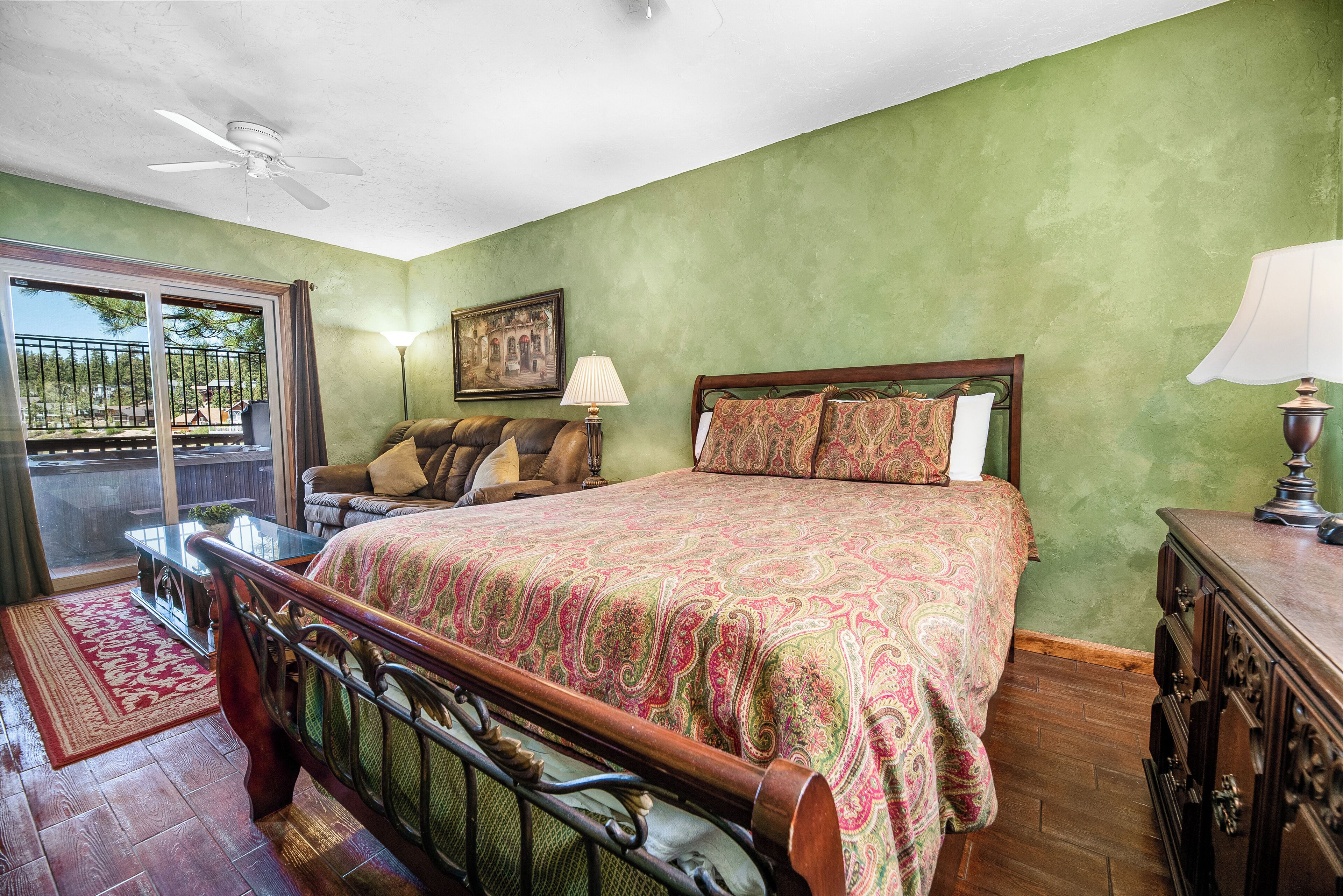 A bedroom with a bed, a ceiling fan, a window with a view, and a nightstand with a lamp.