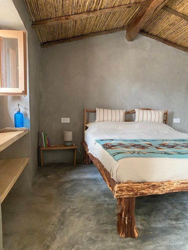 Ein Schlafzimmer mit hölzernem Bettgestell, einem Betonboden, einem kleinen Nachttisch mit Büchern und einer Lampe sowie einem Holzregal.
