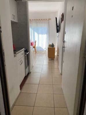 Um corredor estreito com uma cozinha à esquerda, uma área de jantar com mesa e cadeiras no centro, e um pequeno armário de madeira à direita.