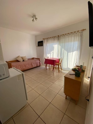 Um quarto pequeno e bem iluminado, com uma cama, uma escrivaninha com computador, uma mesa com cadeiras e uma televisão na parede.
