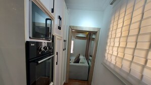 A narrow hallway with a kitchen area on the left, a window with blinds on the right, and a bed in the background.