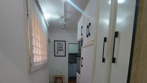 A narrow hallway with wooden flooring, white walls, and a ceiling fan. A window with blinds is on the left, and a kitchen area is visible in the background.
