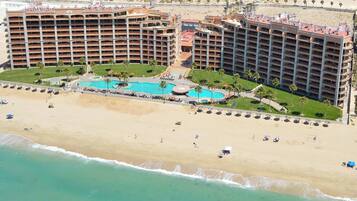 Un grand hôtel à étages, doté d’une piscine centrale et d’un espace plage aménagé avec des parasols et des chaises longues.